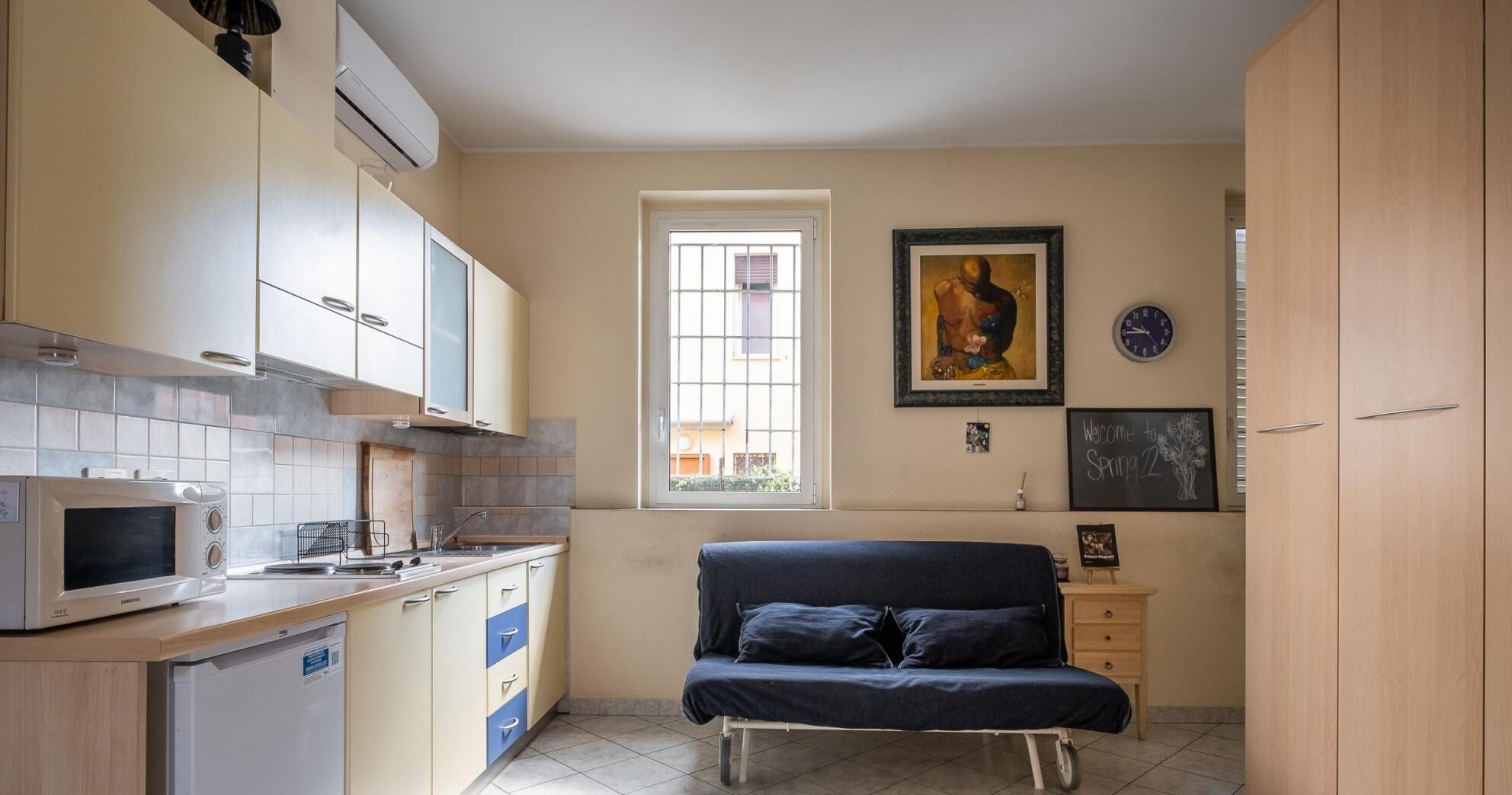 Une petite cuisine équipée d’un four à micro-ondes, d’un évier et de placards. On y trouve une fenêtre avec des barreaux, un canapé et une horloge au mur.