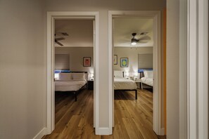 A hallway leading to two bedrooms with wooden flooring, ceiling fans, bedside lamps, and framed pictures on the walls.