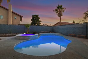 A swimming pool with a hot tub, surrounded by a brick wall, palm trees, and a house.