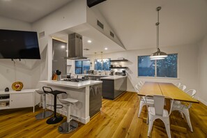 Uma cozinha moderna com uma ilha central, banquetas de bar, uma mesa de jantar com cadeiras e uma TV fixada na parede.
