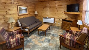 A cozy cabin interior with a wooden ceiling, a ceiling fan, a television mounted on a wall, a sofa, a coffee table, and patterned chairs.