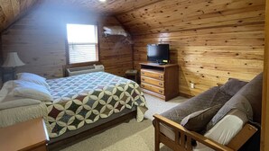 A cozy cabin room with a wooden ceiling, a bed with a patterned quilt, a wooden bench, a dresser, and a television on a stand.