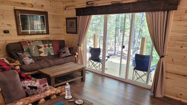 A cozy cabin interior with a wooden floor, a sofa with patterned cushions, a coffee table, and a large sliding glass door leading to an outdoor deck.