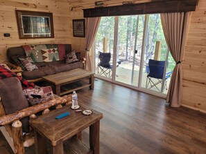Interior acollidor d'una cabana amb sòl de fusta, un sofà amb coixinets estampats, una taula de cafè i una gran porta corredissa de vidre que dona a una terrassa exterior.