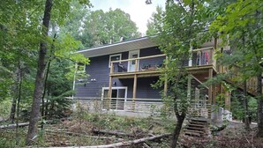 A modern house with a balcony and wooden deck, surrounded by trees and overgrown vegetation.