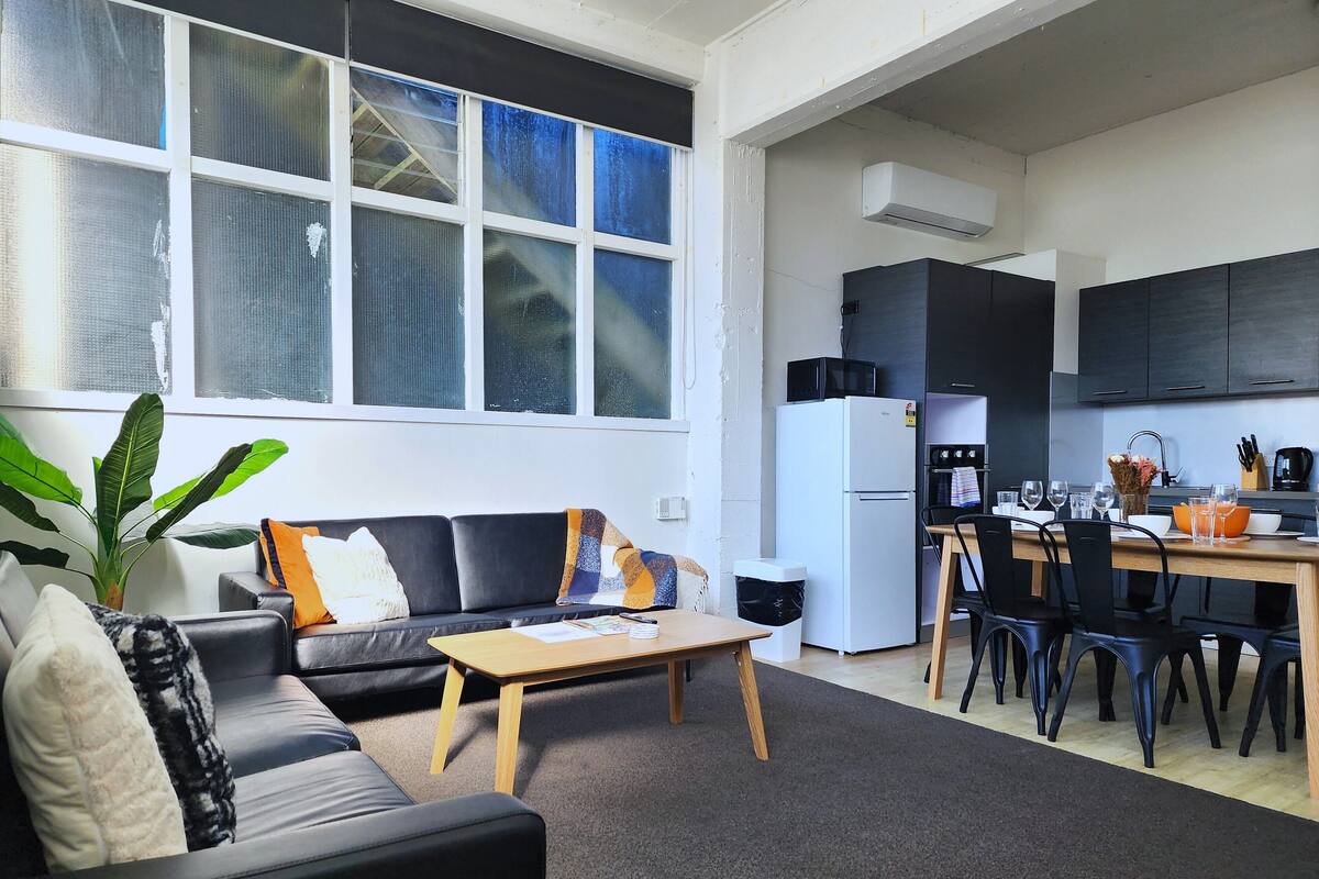 A modern living room with a black sofa, a wooden coffee table, and a large window with blinds.