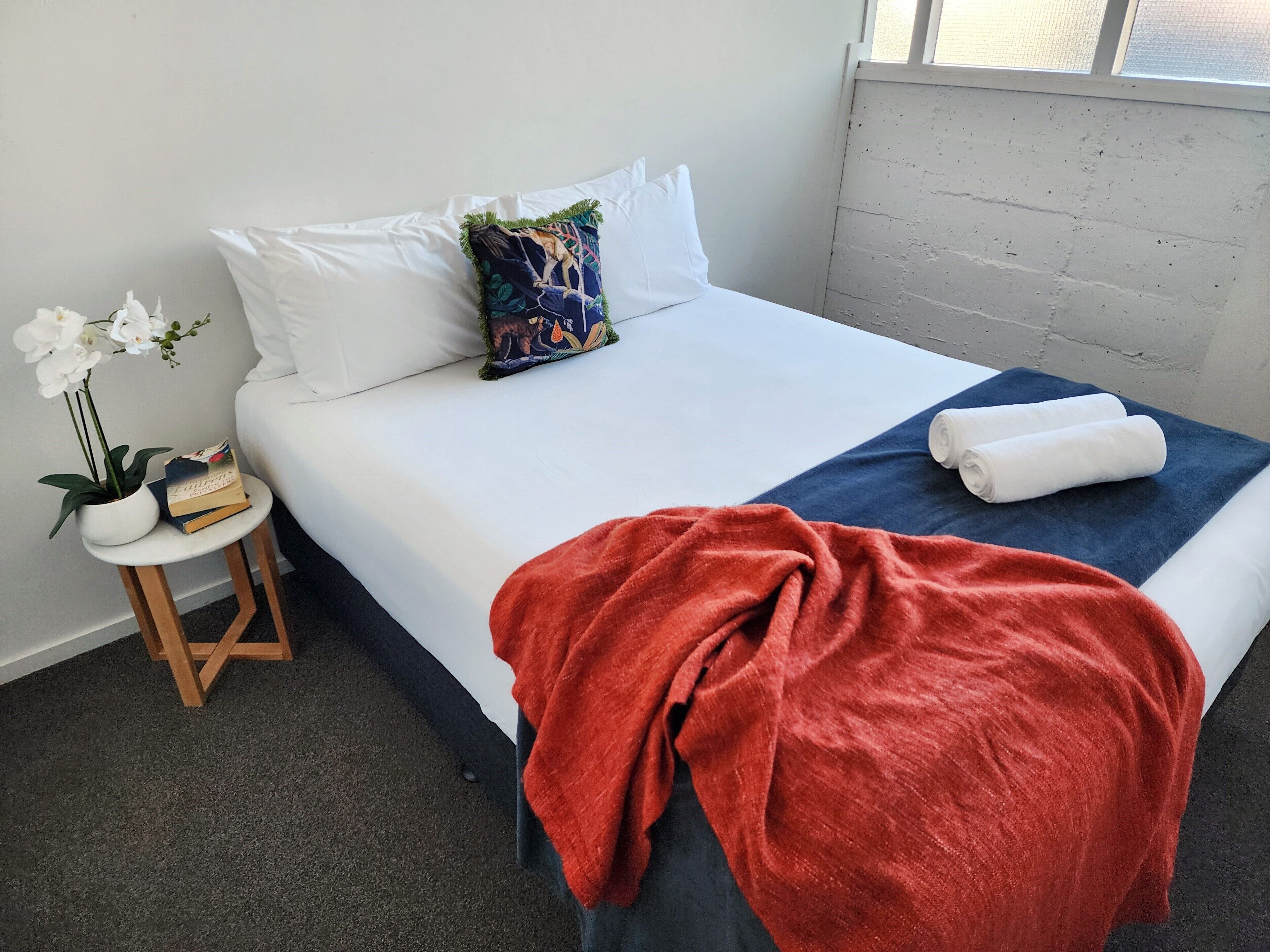 A neatly made bed with white and blue bedding, a red throw blanket, and a decorative pillow. A small wooden side table with a potted plant and a book is beside the bed.