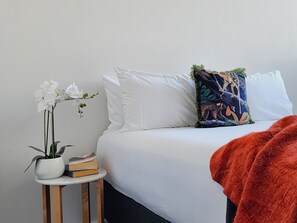 Una cama bien hecha con ropa de cama blanca, una mesita de noche con una planta en maceta y libros, y un cojín colorido.