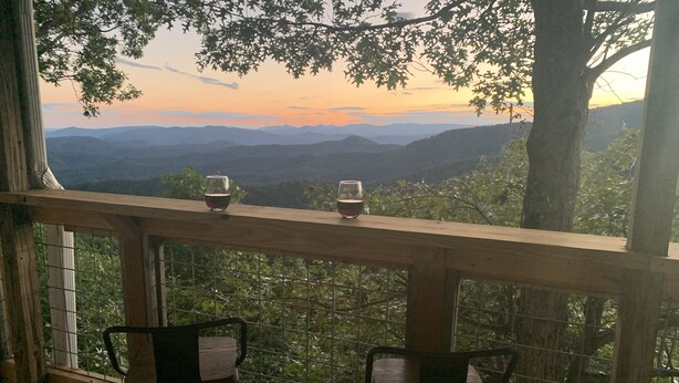 Um deck de madeira com duas cadeiras voltadas para uma vista panorâmica de montanhas e um pôr do sol.