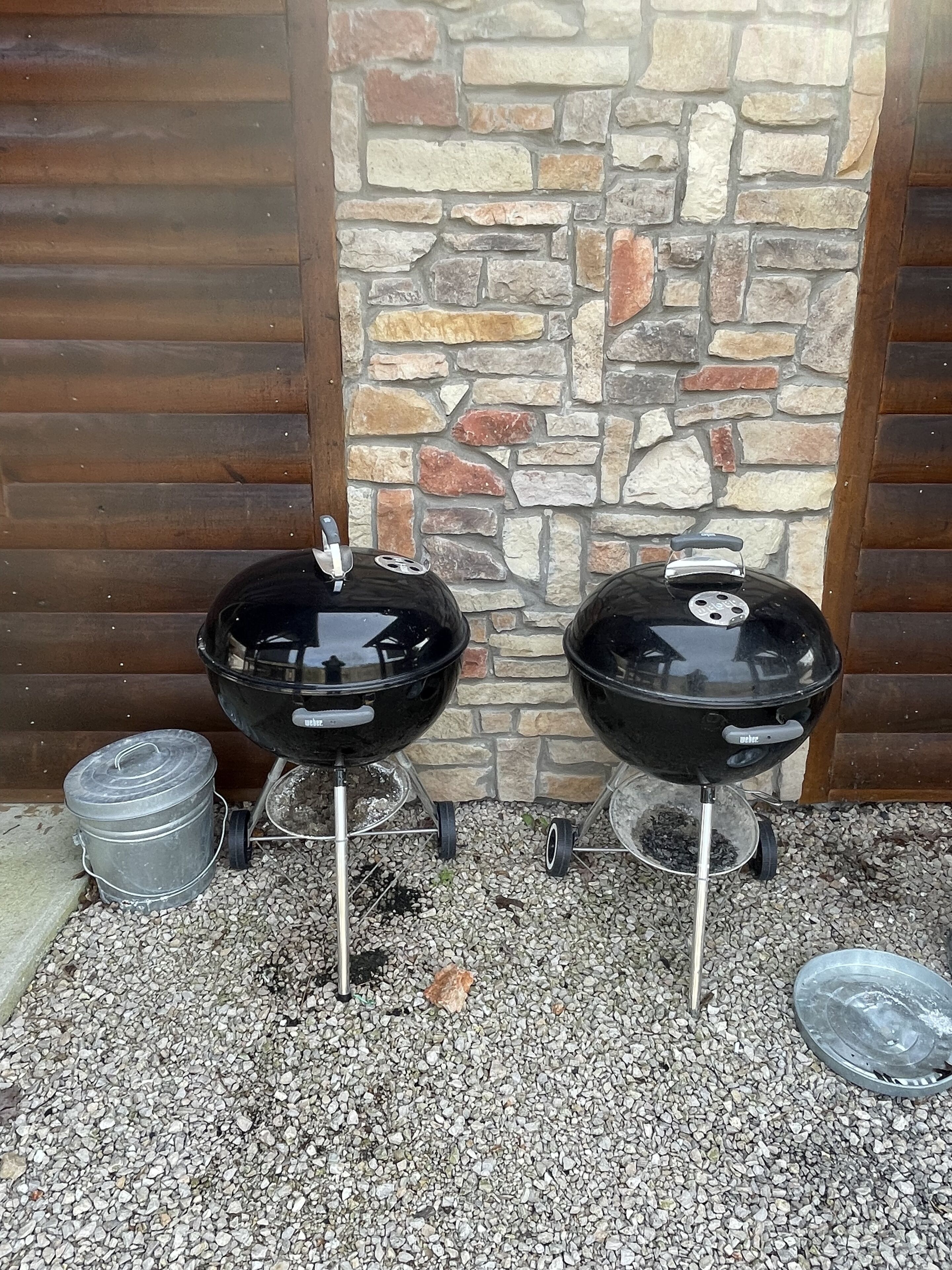 Two black barbecue grills with wheels, a stone wall, and a wooden door in the background.