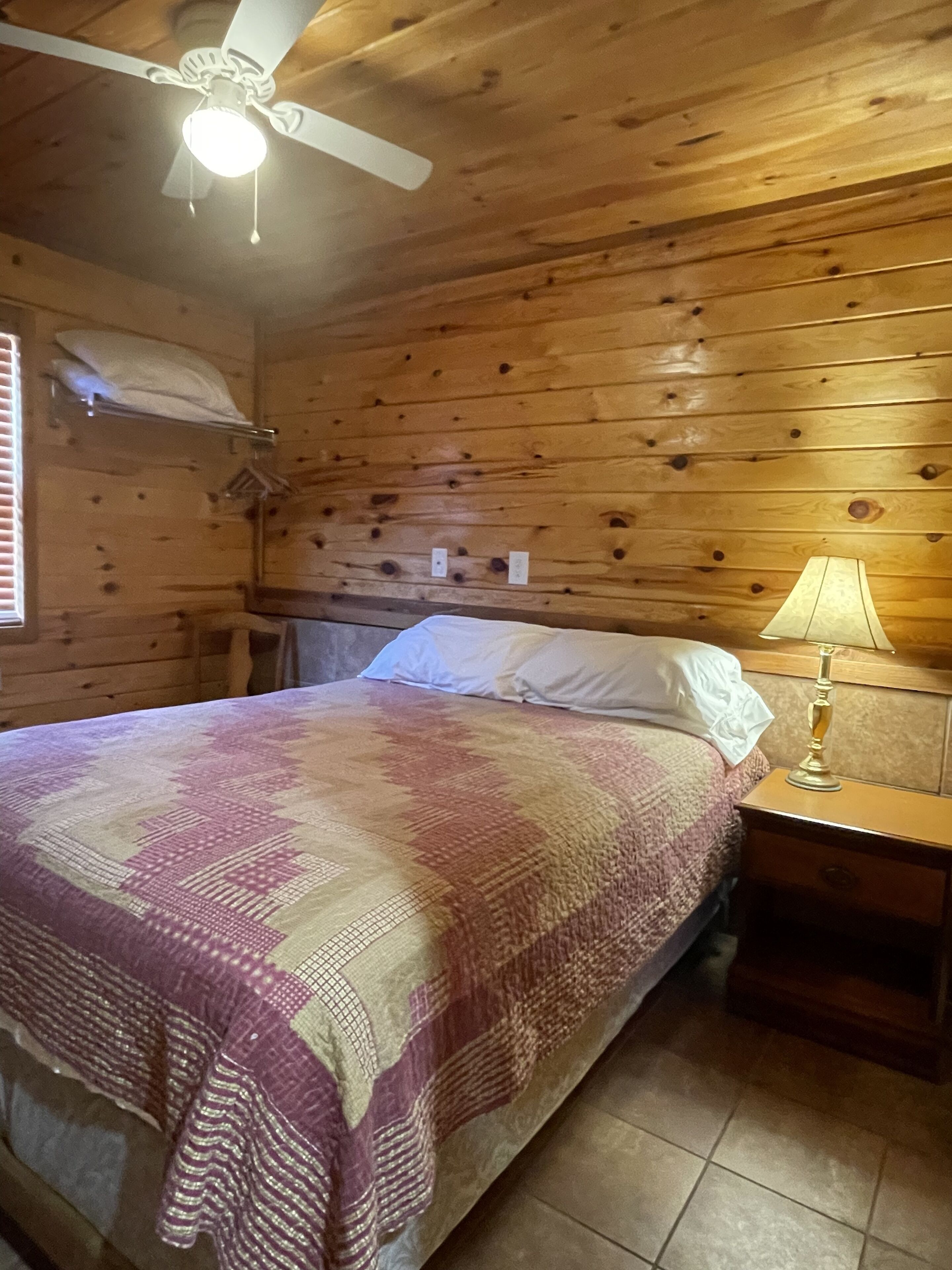 A wooden cabin room with a bed, a nightstand, a lamp, and a ceiling fan.