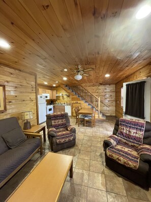Interior acogedor de una cabaña con techo de madera, comedor, sofá y cocina al fondo.