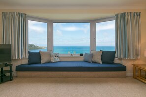 A room with a bay window overlooking the sea, a sofa with cushions, a TV stand, and a lamp. - Dolphin Watch (St Ives)
