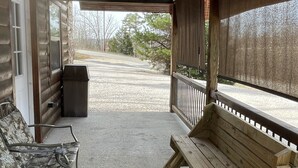 Un porche abrité avec un banc en bois et une chaise à motifs, un sol en béton et une vue sur une route et des arbres.