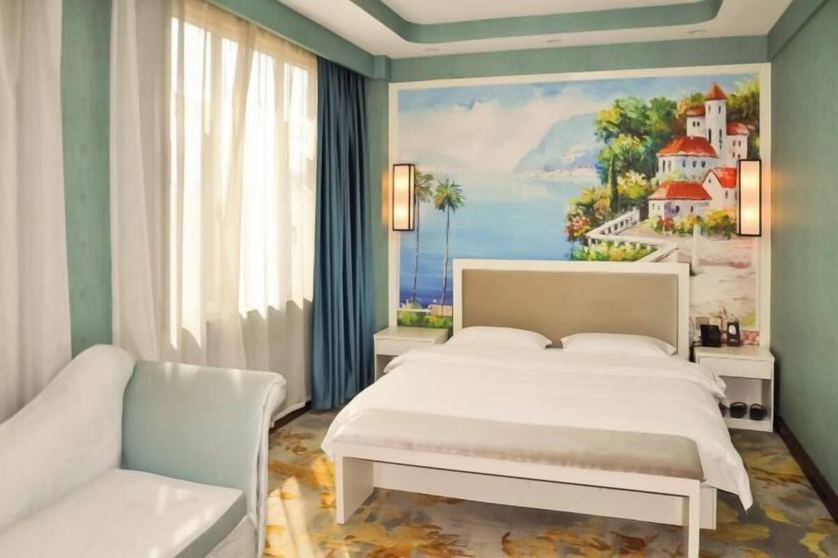 A hotel room with a bed, a chair, a painting of a coastal scene, and a nightstand with a lamp.