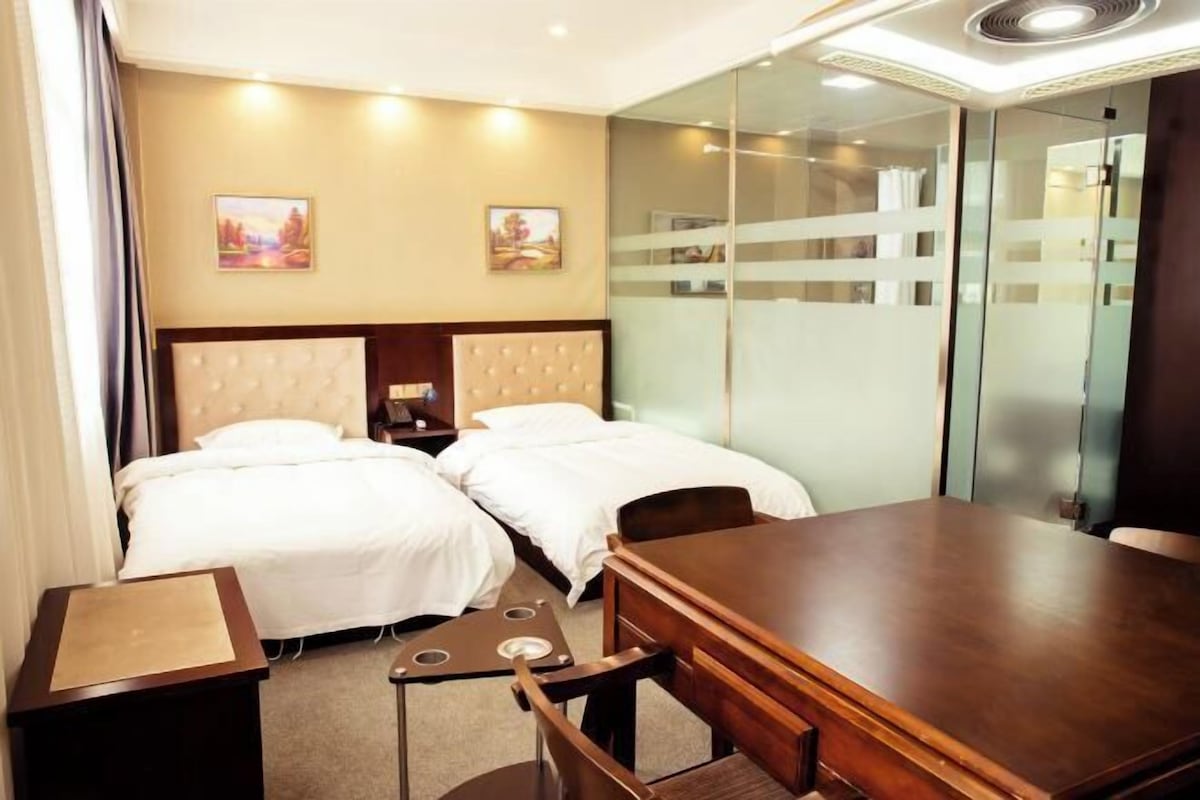 Une chambre d’hôtel avec deux lits, une table en bois, un petit bureau et une cloison en verre.