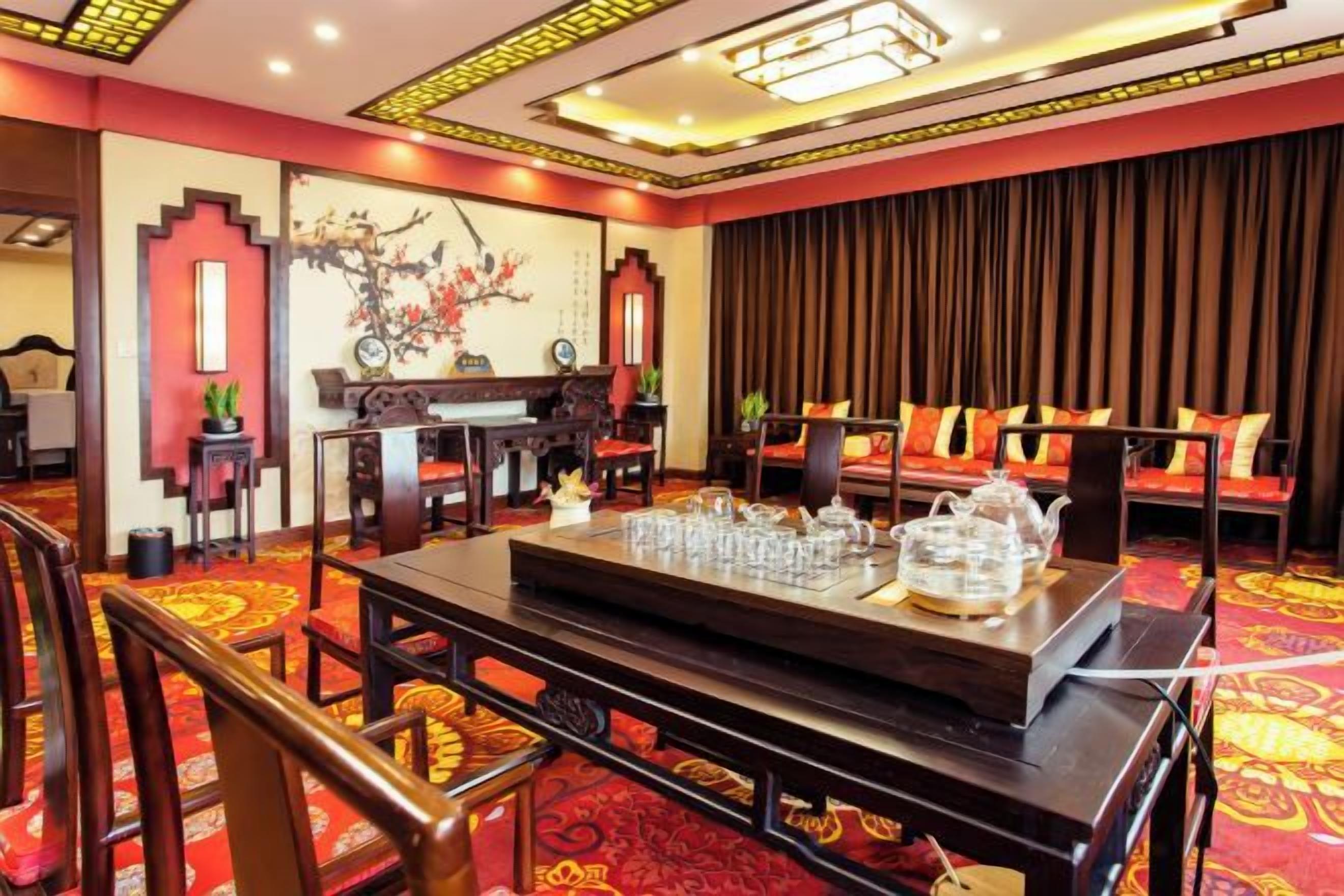 A hotel room with a traditional Asian-style interior, including a dining table set with teapots and glasses, a piano, and a sofa with cushions.