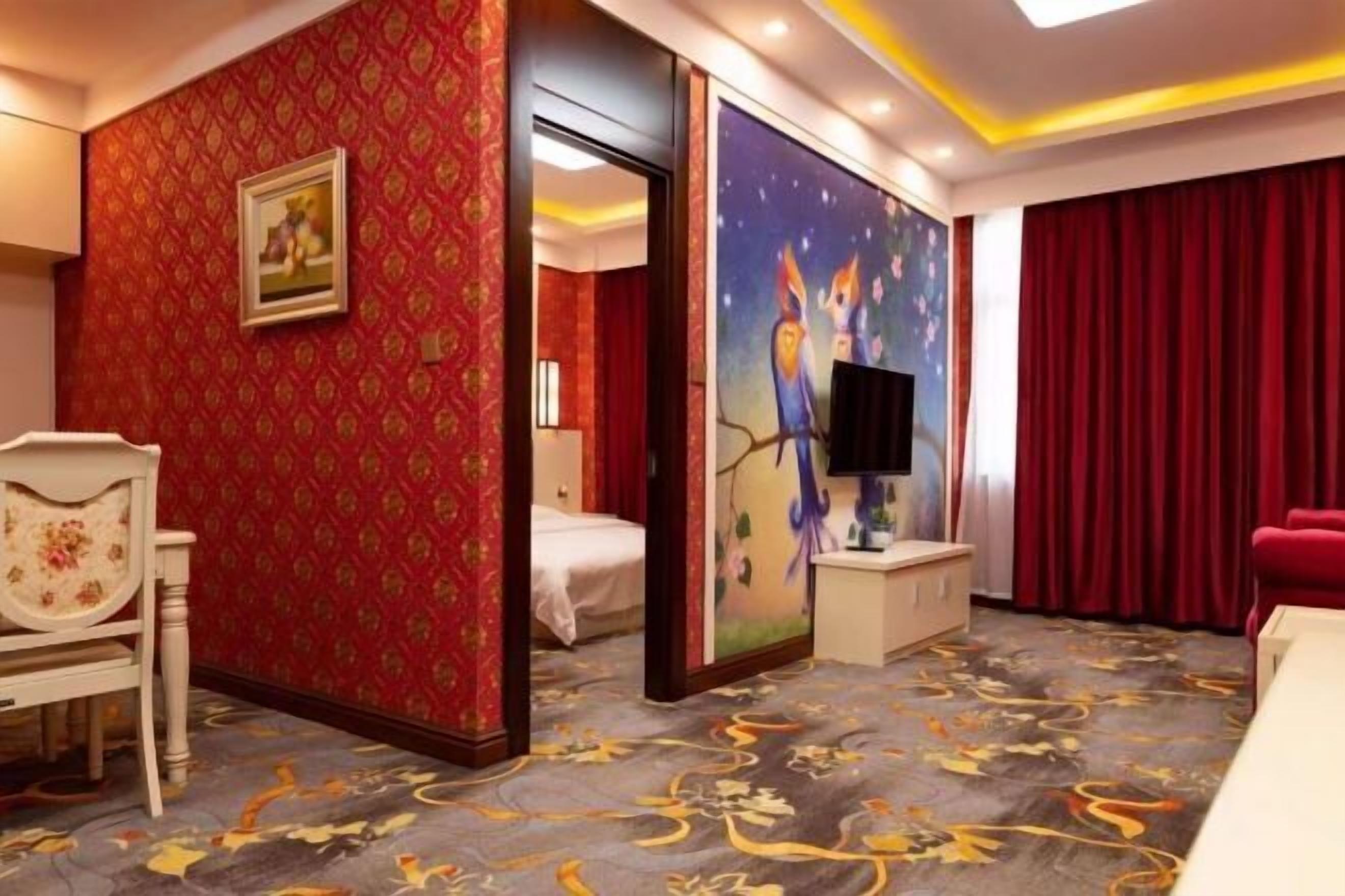 A hotel room with a red patterned wall, a bed, a TV, a desk, and a red curtain.