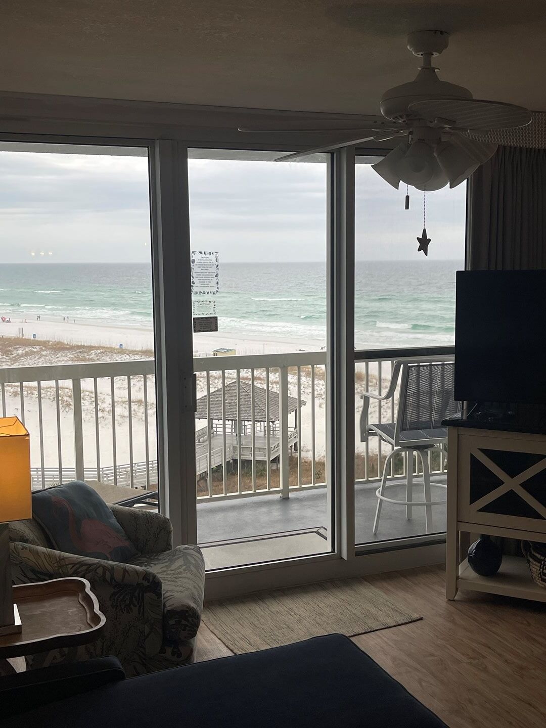 A room with a large window overlooking a beach, a ceiling fan, and a TV on a stand.