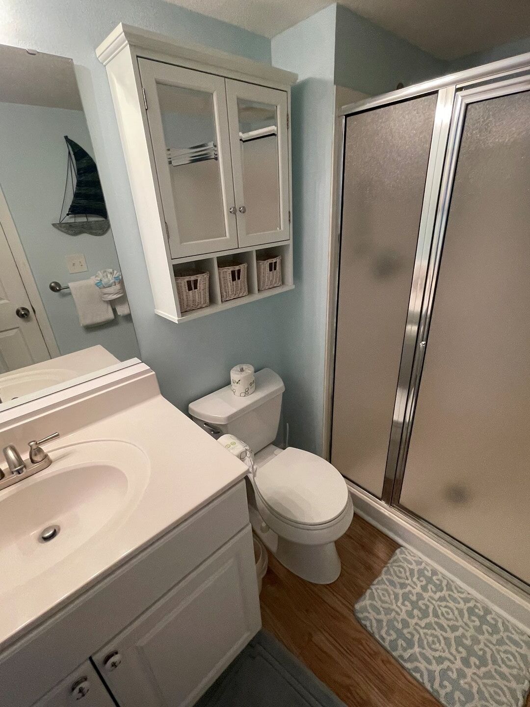 A bathroom with a white sink, toilet, and shower. There are wicker baskets on the wall and a patterned rug on the floor.
