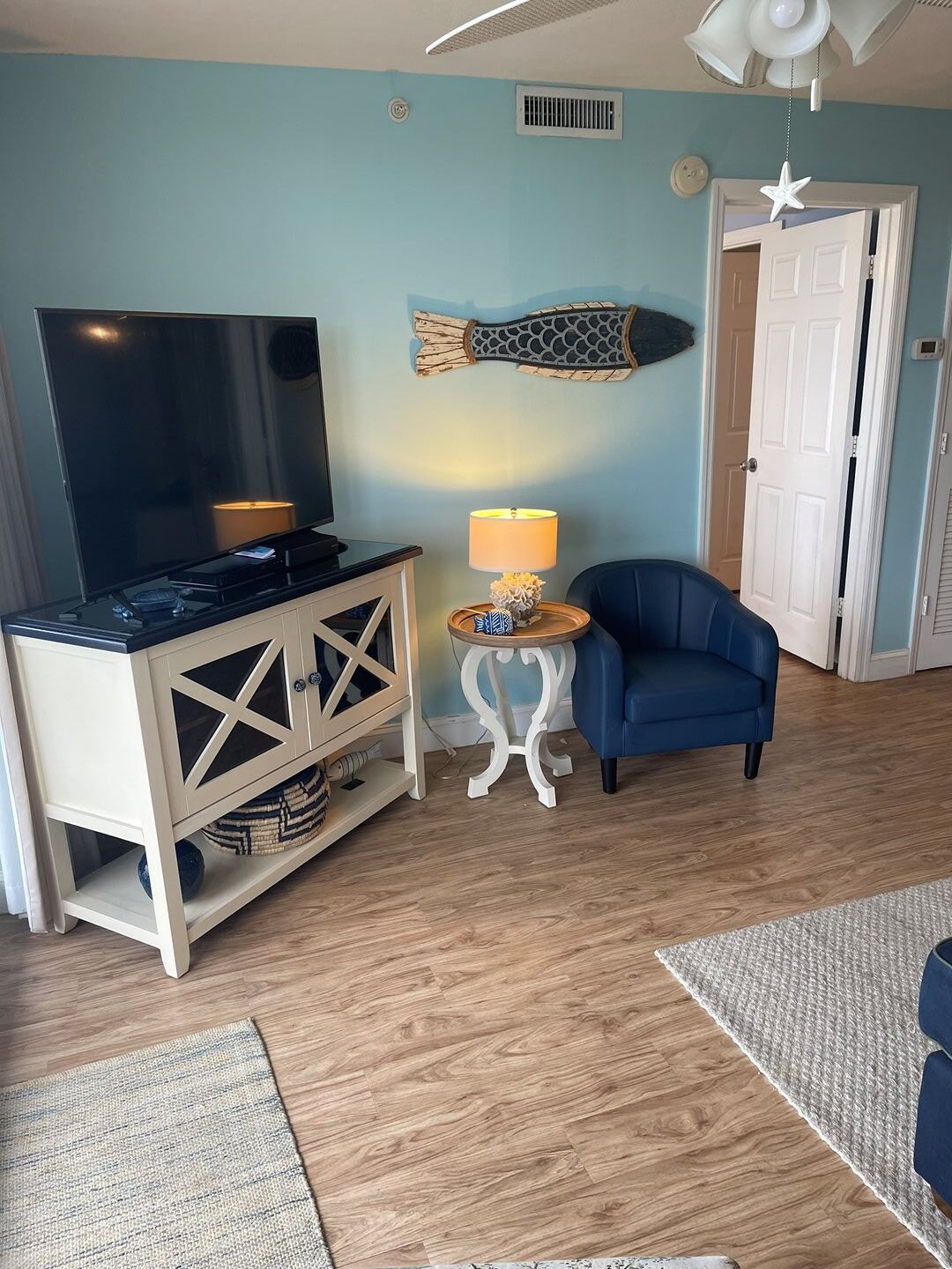 Una sala de estar con televisor, un sillón azul, una mesita con lámpara y un adorno en la pared.