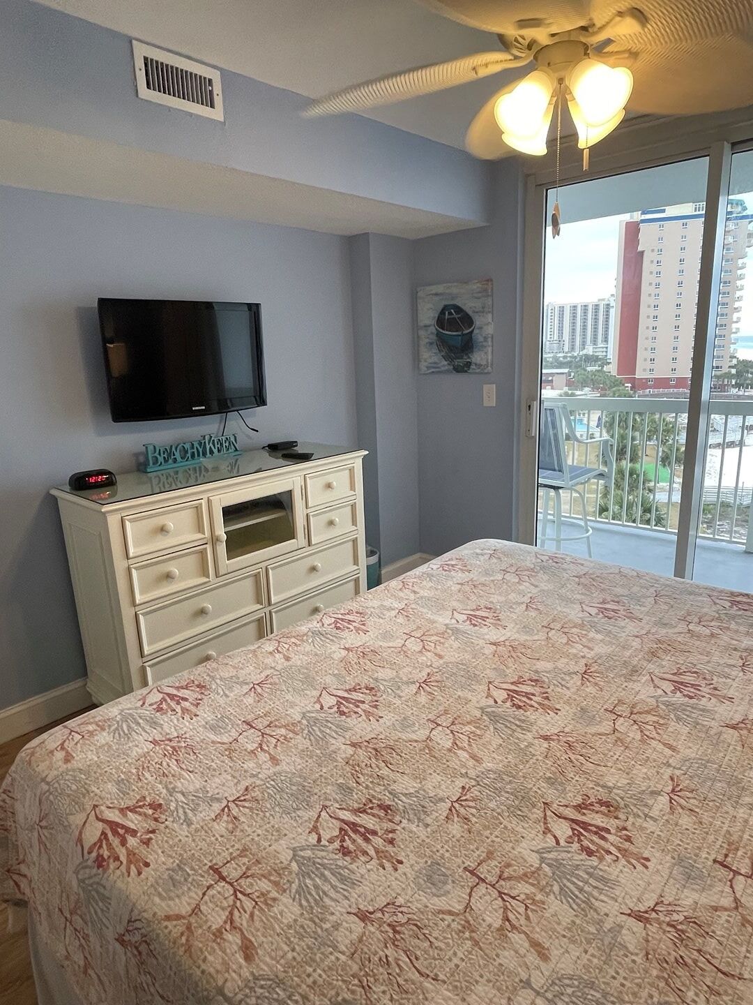 Un dormitorio con cama, cómoda, televisor y vista a la ciudad desde un balcón.