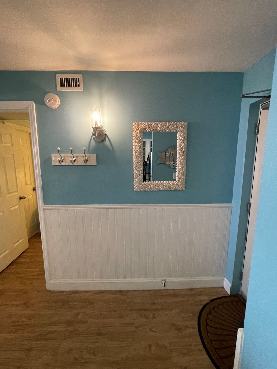 A room with teal walls, a white wainscoting, a wooden floor, a white door, a wall-mounted light fixture, a mirror with a textured frame, and a vent on the ceiling.