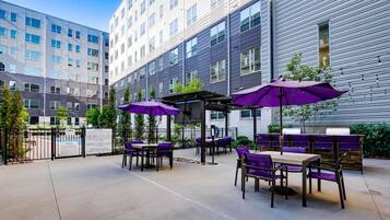 An outdoor patio area with tables and chairs, some with purple umbrellas, and a building in the background.