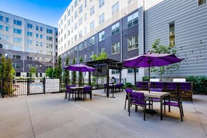 An outdoor patio area with tables and chairs, some with purple umbrellas, and a building in the background.