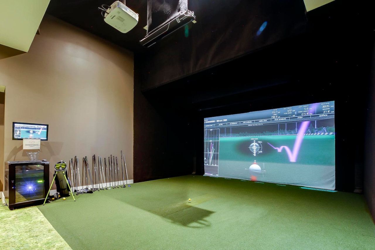 Ein Raum mit einem großen Bildschirm, der eine Golfsimulation zeigt, einem Projektor und einer Golfbag.