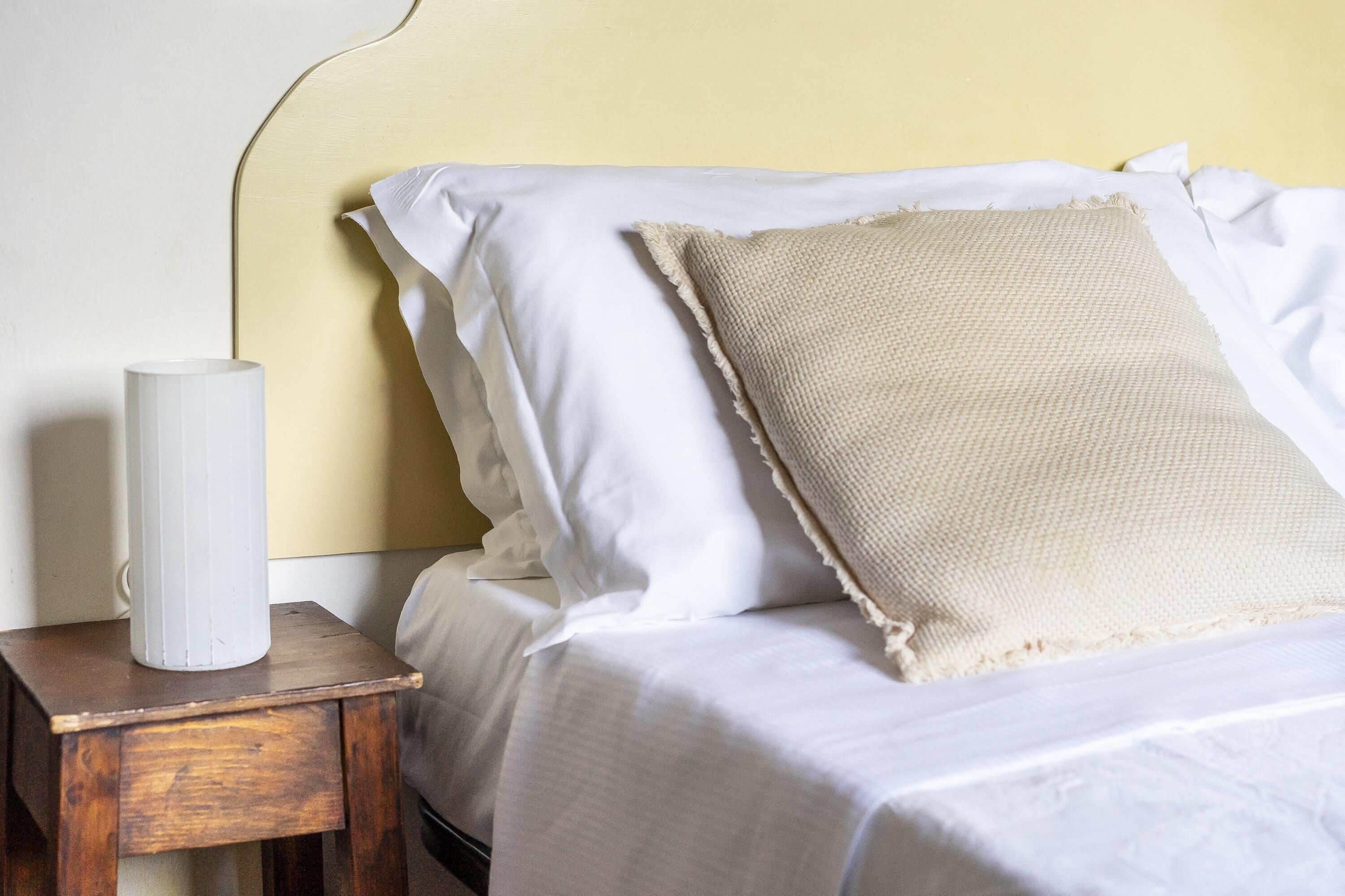 A neatly made bed with white linens and a beige pillow, a white vase on a wooden nightstand beside it, and a yellow wall in the background.