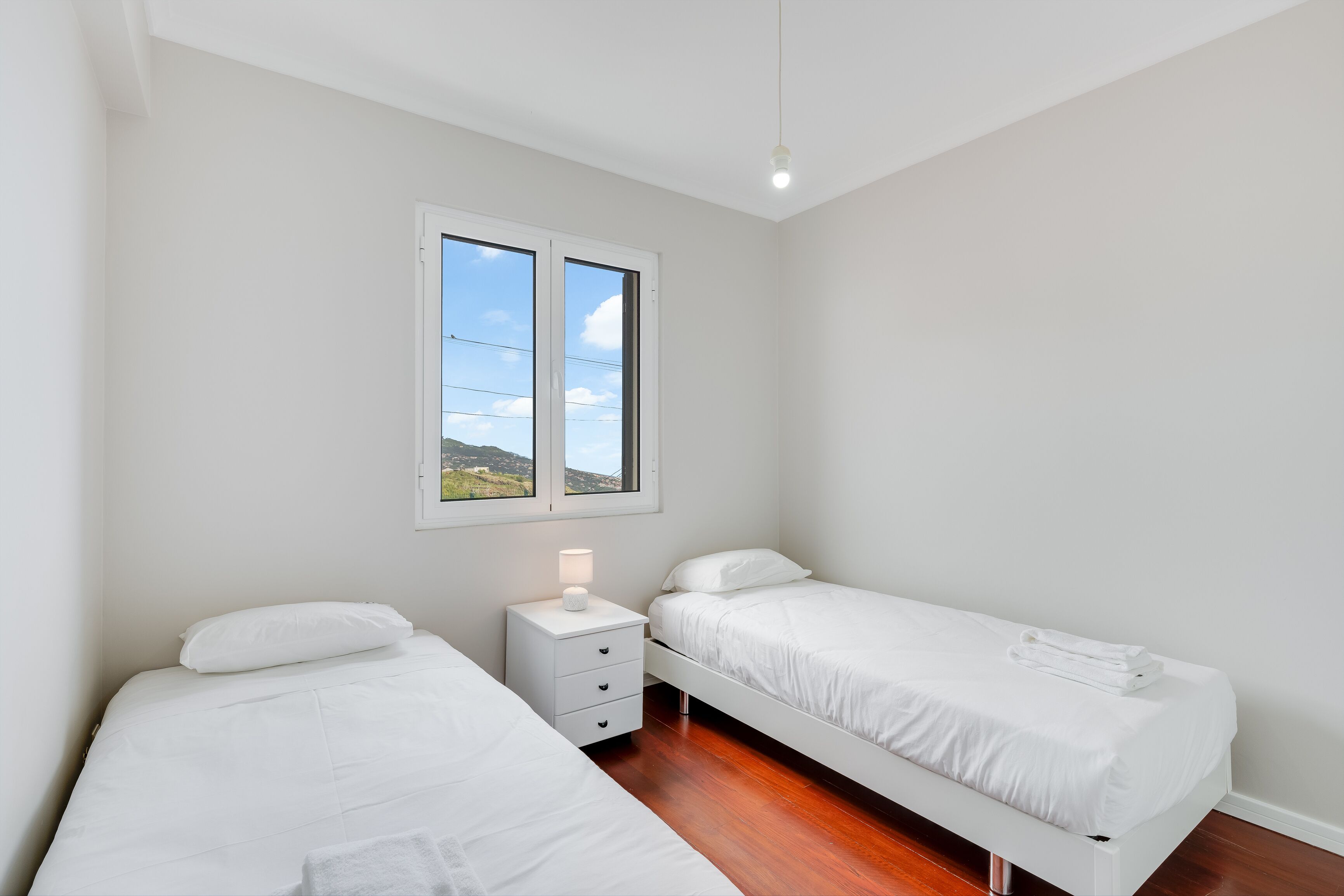 A bedroom with two single beds, a nightstand with a lamp, and a window with a view of the outdoors.