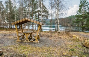Un refugio de madera para picnic con bancos, rodeado por un bosque y un lago.