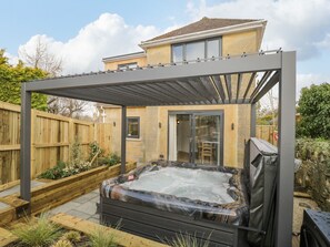 Un jacuzzi bajo una pérgola en un patio trasero con cerco de madera y una casa al fondo.
