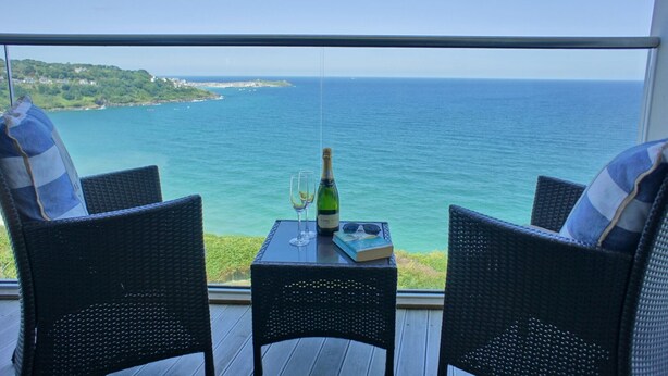 A balcony with wicker chairs, a table with a bottle of champagne, glasses, and a book. The view includes a coastline and the sea. - Glas Mor (St Ives)