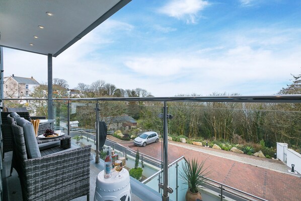 A balcony with wicker furniture, a glass railing, and a view of a street with a car. - Bamaluz (St Ives)