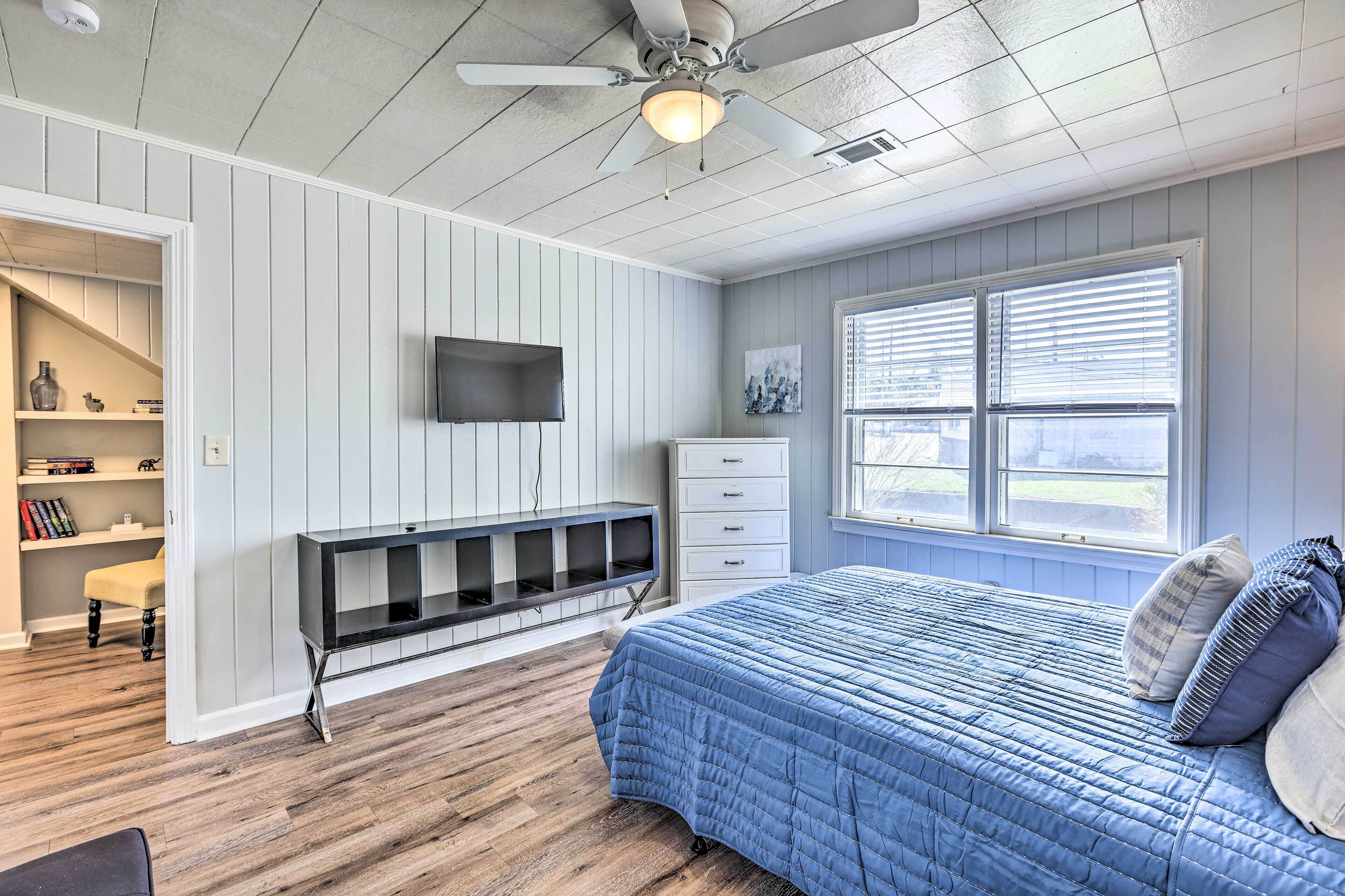 A bedroom with a bed, a TV mounted on the wall, a ceiling fan, and a window with blinds.