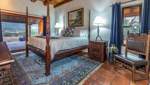 Un dormitori amb un llit amb cortines, dues taules de nit amb làmpades, una cadira, un finestral gran amb cortines i un quadre a la paret.