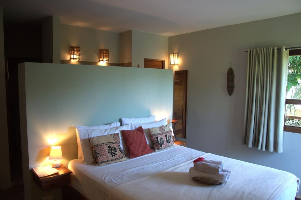 A neatly made bed with white linens and patterned pillows, a bedside lamp, and a window with curtains.