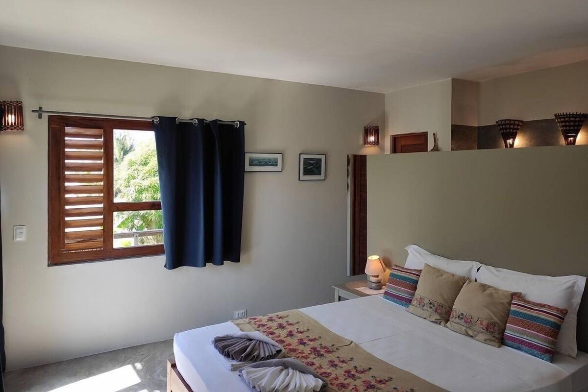 A bedroom with a bed, a window with curtains, a bedside table with a lamp, and a wall-mounted shelf with decorative items.