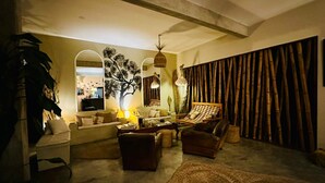 Uma sala de estar aconchegante com um sofá, poltronas, uma mesa de centro e um tapete grande. Há uma parede com um mural de uma árvore e uma cortina de bambu.