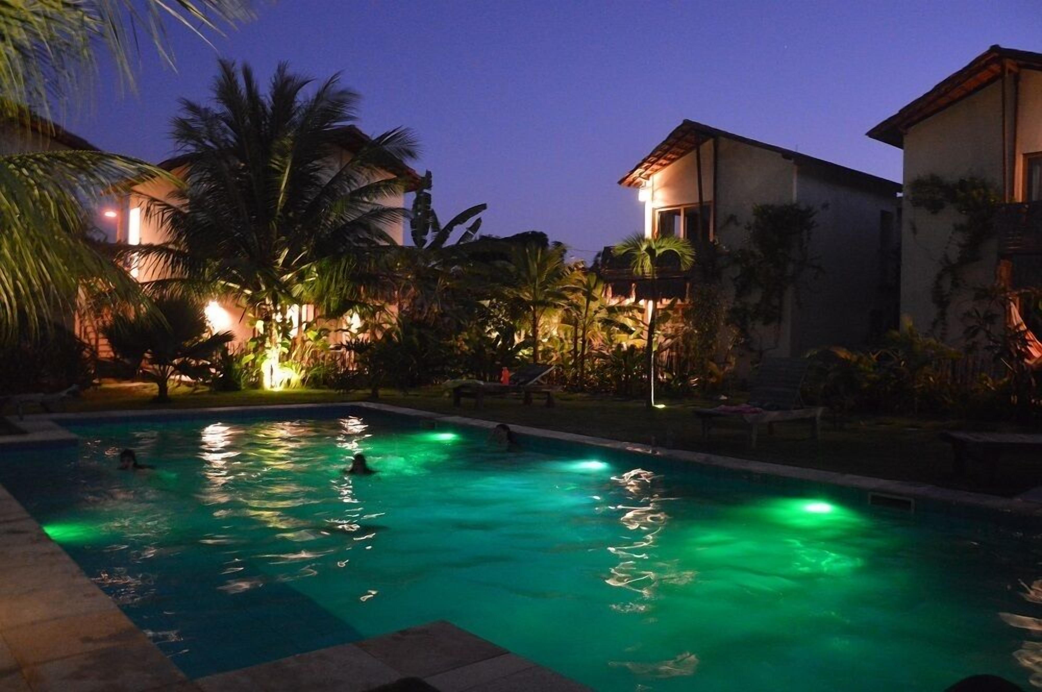 Piscina com pessoas nadando, rodeada por palmeiras e edifícios ao entardecer.