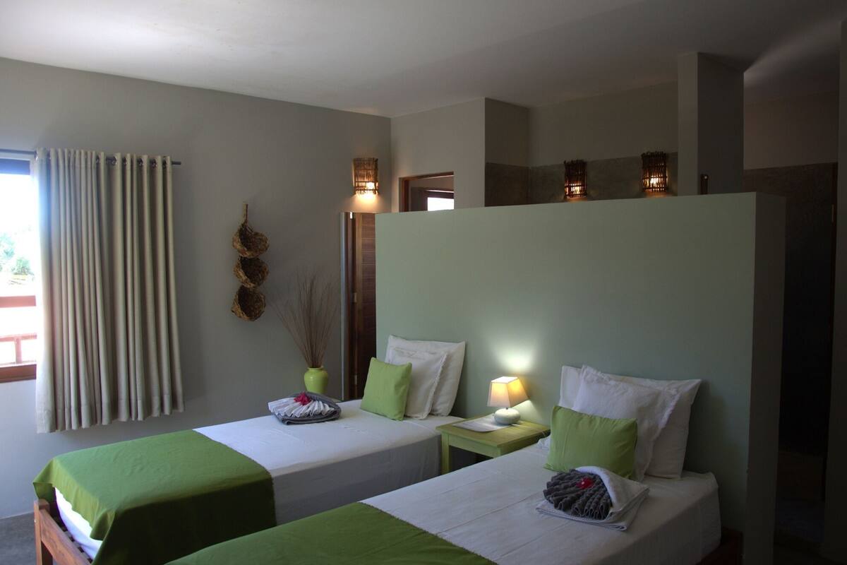 A hotel room with two beds, a window with curtains, a wall-mounted light fixture, and a small table with a lamp.