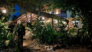 Um jardim tropical com uma construção de palha, cercado por vegetação exuberante e um caminho de terra.
