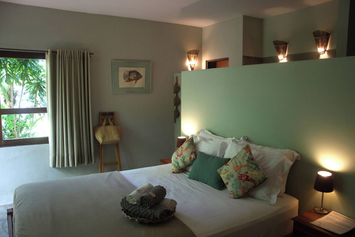 A neatly made bed with pillows and a rolled towel, a small chair, and a wall-mounted lamp.