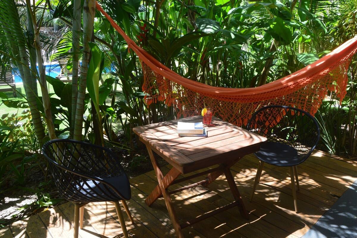 A wooden deck with a hammock, two chairs, and a table with books and a drink.