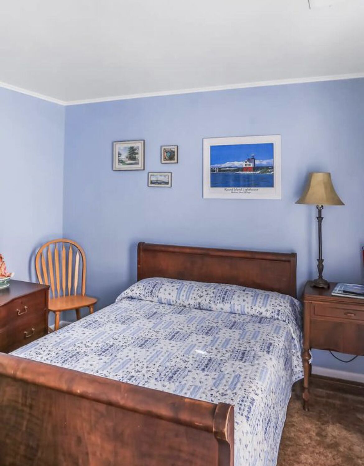 A bedroom with a bed, wooden nightstand, chair, and a lamp. There are three framed pictures on the wall.