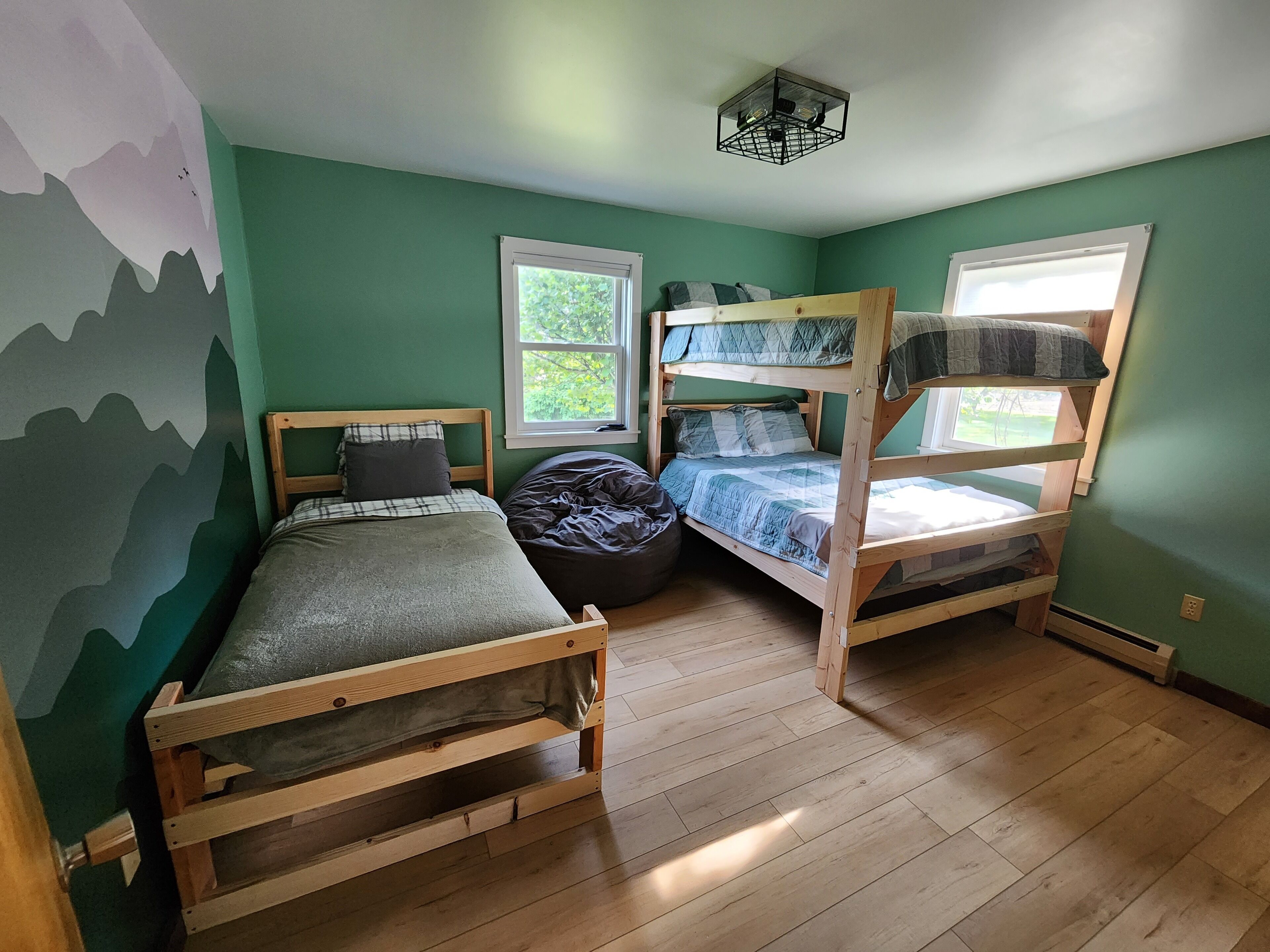 Une chambre avec deux lits superposés, un lit simple, un fauteuil poire et une fenêtre donnant sur un paysage verdoyant.