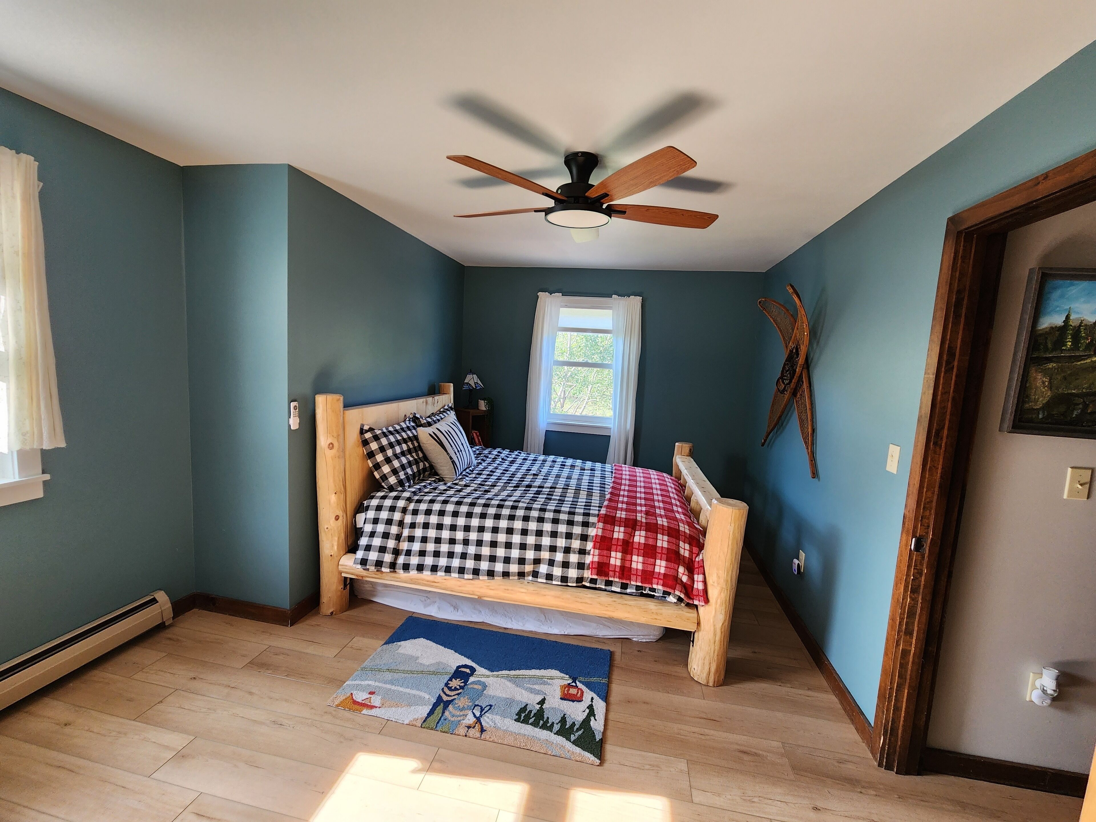 Quarto com cama de madeira, ventilador de teto, janela com cortinas, um adorno na parede e uma porta.