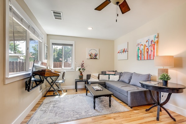A living room with a sofa, coffee table, and a desk with a chair. There are paintings on the wall and a window with blinds.